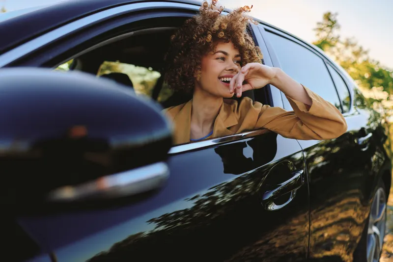 A woman driver leans from a sleek car, smiling with relaxed confidence. Bright outdoor light and retro color tones create a bold lifestyle moment, emphasizing candid joy and movement.