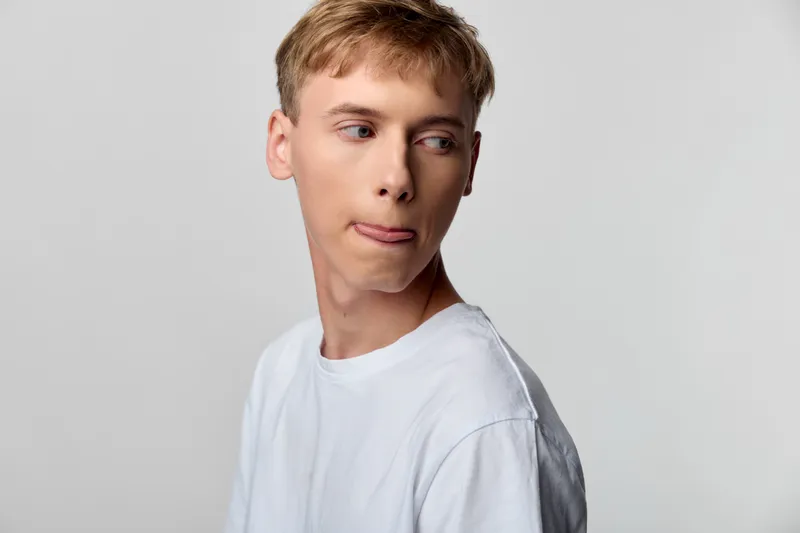 Casual man in white t shirt, side profile with playful tongue out, simple studio backdrop, relaxed mood and expressive gaze, modern stock photography suited for lifestyle or youth campaigns