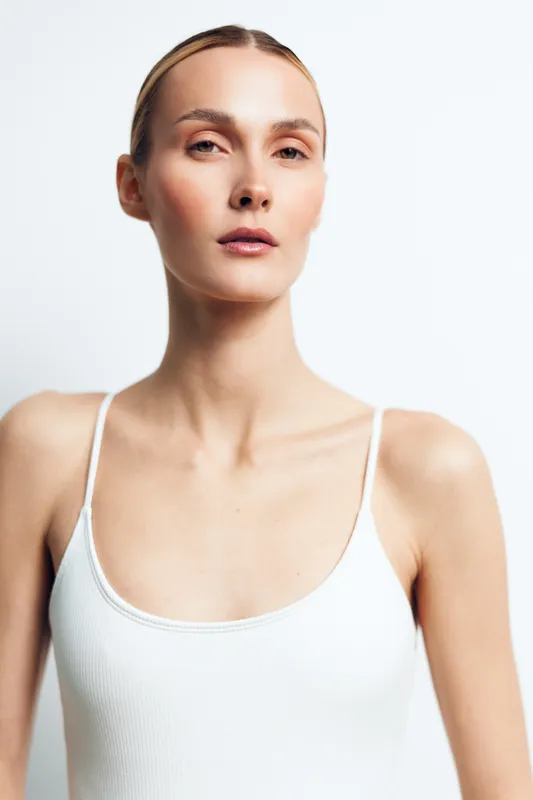 Portrait of confident woman with natural skin, wearing white tank top, minimal makeup, clean background, soft lighting, elegant expression, simple hairstyle, fresh and healthy look