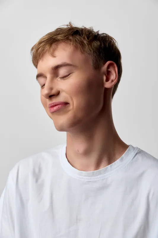 Relaxed, smiling male in a white casual tee in a studio setting with soft lighting, closed eyes, serene mood, minimal fashion vibe, clean background and focus on calm expression
