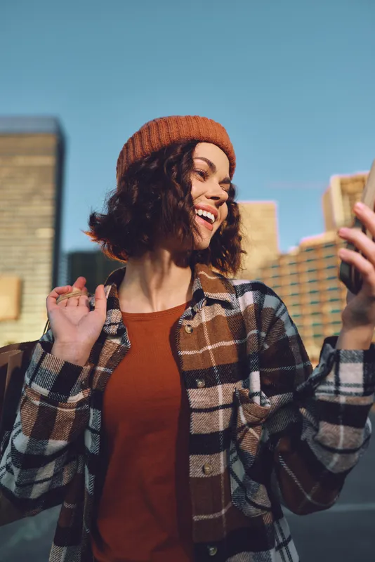 Woman smile with phone in urban setting, candid beanie and jacket portrait in sunlight showing authenticity, candid lifestyle, golden hour glow, mindful living and emotional storytelling.