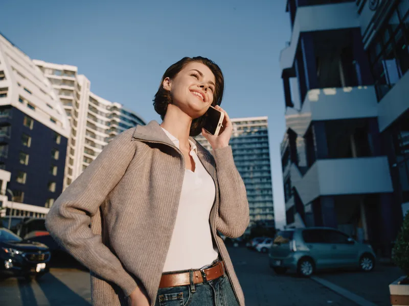 A cheerful professional woman stands outdoors in an urban setting, talking on a smartphone with a confident smile. Her relaxed pose and bright expression convey a positive, modern business atmosphere.
