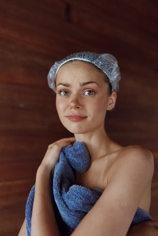 young woman in shower cap wrapped in towel with calm expression indoors, soft natural light and wooden background creating cozy atmosphere, skin care routine concept
