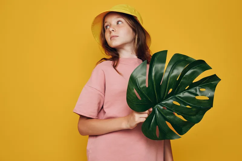 Teenage girl in a pink shirt and yellow hat holding a large green tropical leaf on a yellow background. Studio portrait with natural concept and summer vibe.