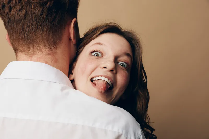Playful woman with brown hair sticks out tongue while hugging man in white shirt. Close-up on beige background. Concept of fun, affection, and lighthearted relationship moments.