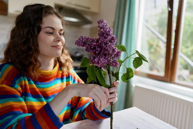 Cheerful woman indoors arranging a bouquet, colorful striped sweater, soft sunlight from a window, relaxed mood, cozy home setting, vibrant lilac flowers, everyday lifestyle