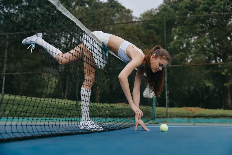 Young athletic woman preparing to serve a tennis ball, wearing sporty attire on a court surrounded by greenery, expressing focus and determination in her pose