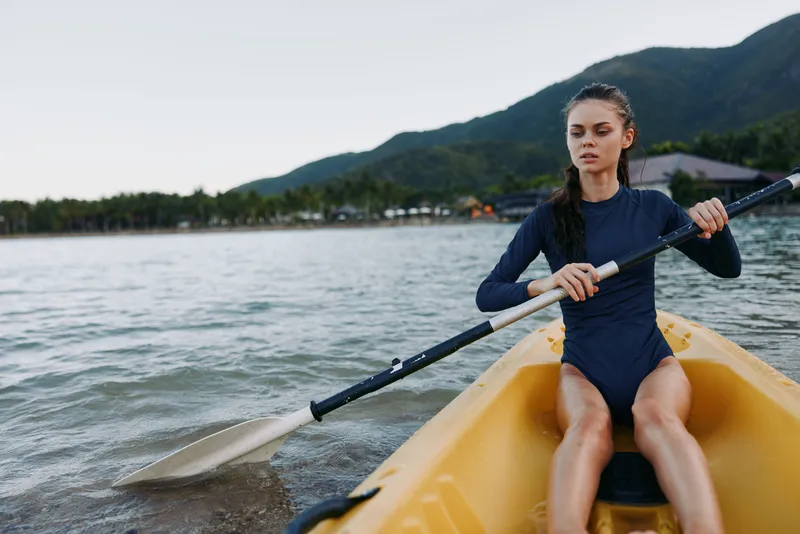 Female kayaker paddling a yellow kayak on calm water, wearing a dark wetsuit, focused expression as she grips a paddle mid stroke.