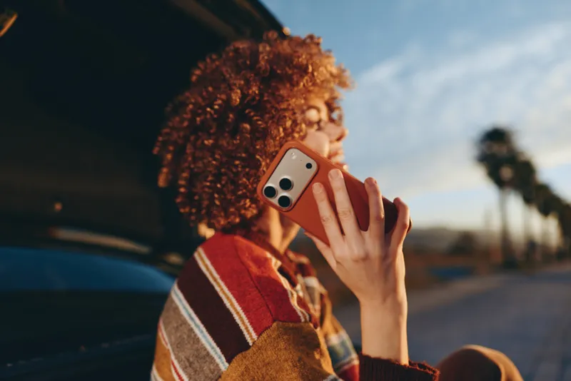 Woman lifestyle outdoors, curly hair woman holding phone on street, smiling softly, warm evening light, casual sweater, relaxed posture, urban background, natural colors, candid moment