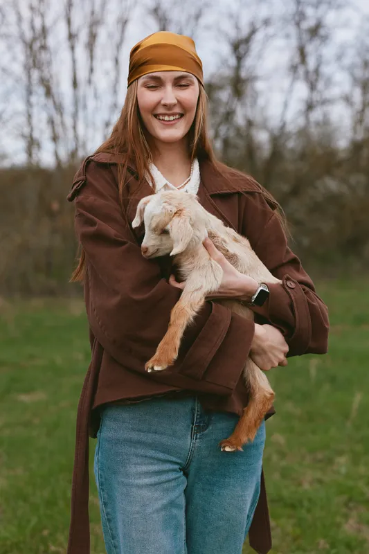 Happy woman outdoors in casual clothing holds a goat kid in a park, creating a wholesome lifestyle moment with affection and companionship under natural lighting in nature.