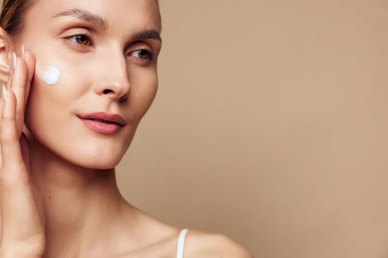 Young woman applying moisturizer on face, gentle skincare routine, natural makeup, soft lighting, smooth skin texture, beige background, calm expression