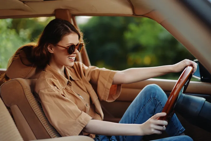 Young woman driving a car, wearing sunglasses and a beige oversized shirt, smiling with joy, showcasing a carefree lifestyle in a natural outdoor setting.