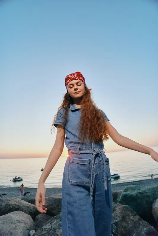 lifestyle woman in modern boho-western denim street style with film color effects, standing outdoors near the seaside during sunset, youthful and relaxed