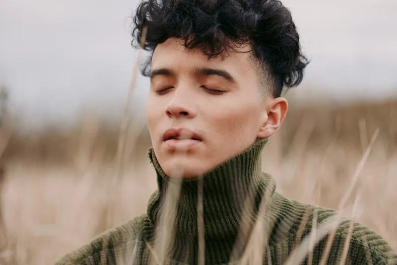 Portrait of a young person with eyes closed, wearing a green turtleneck, calm expression, and a natural field backdrop. An authentic, candid moment captured with soft light.