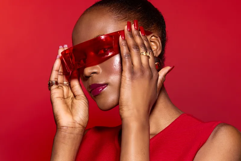 Stylish dark skinned African woman with short hair wears red sunglasses and red dress against red background. Fashion portrait with glossy lips and manicured nails. Concept of modern beauty.