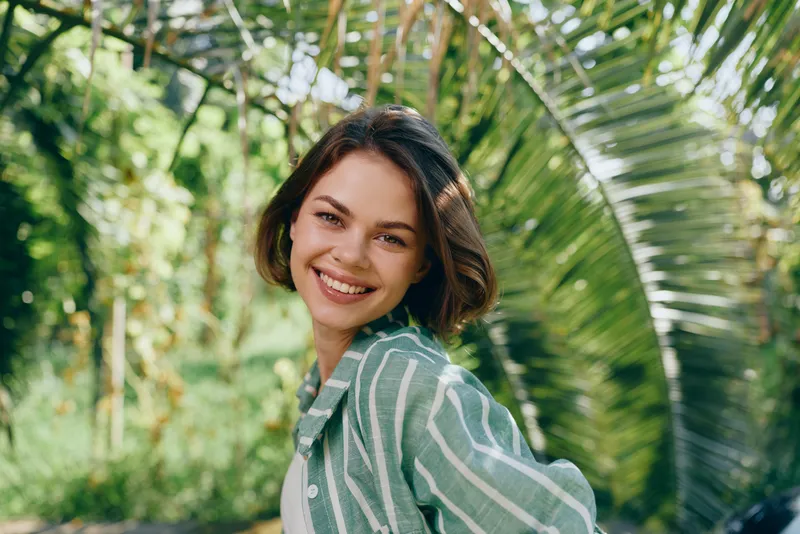 Portrait of a smiling woman outdoors, wearing a green striped shirt, with lush greenery and soft natural light in the background.