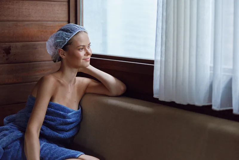 Relaxed woman wrapped in blue towel and wearing hair cap, sitting by window with soft natural light and calm expression, indoor wellness and spa atmosphere.