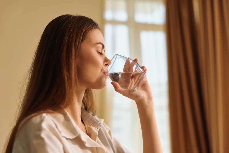Woman glass water drinking at home in morning, relaxed portrait of healthy adult female sipping clear water from tumbler by window with natural light and calm wellness routine.