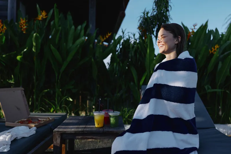 Smiling woman wrapped in striped towel sitting outdoors near table with drinks, morning sunlight, relaxed mood, tropical plants in background, casual lifestyle moment