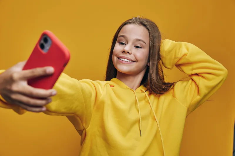 Girl in yellow hoodie taking selfie with red phone on yellow background smiling with hand behind head, casual portrait with happy expression, social media content creation.