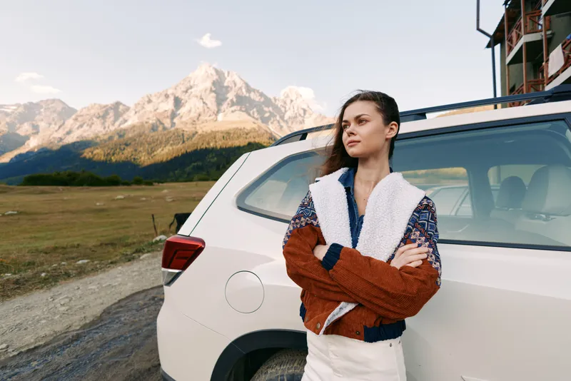 Woman leaning against white car in mountains landscape, portrait of a confident traveler on a roadtrip. Travel adventure outdoors scene with warm jacket, arms crossed and serene scenery.