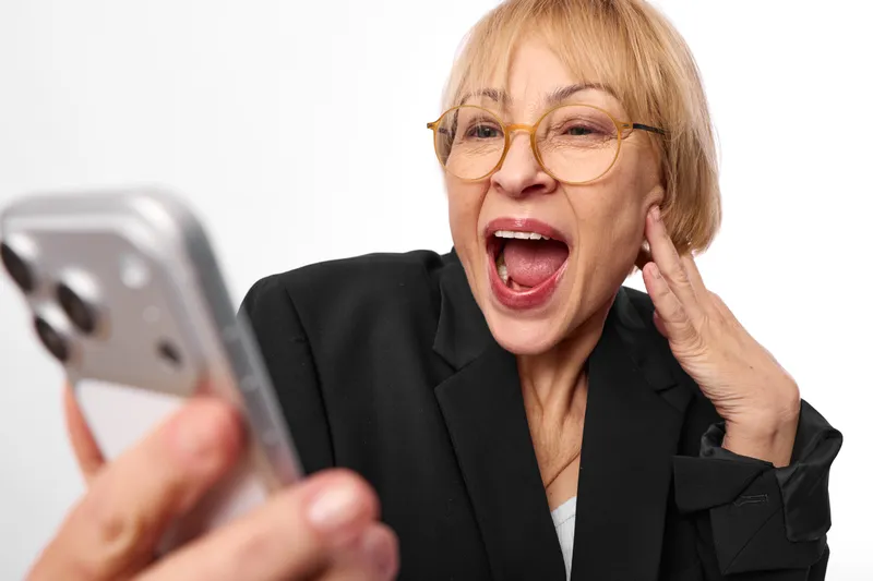 Elderly woman with glasses in business jacket holds smartphone and shouts in surprised emotion, hand near cheek, close up portrait on white studio background