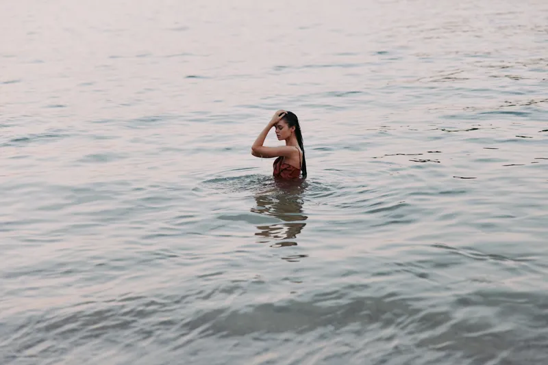 A solitary swimmer stands waist-deep in calm water, shielding eyes with a hand as soft light highlights ripples and creates a serene seaside moment.