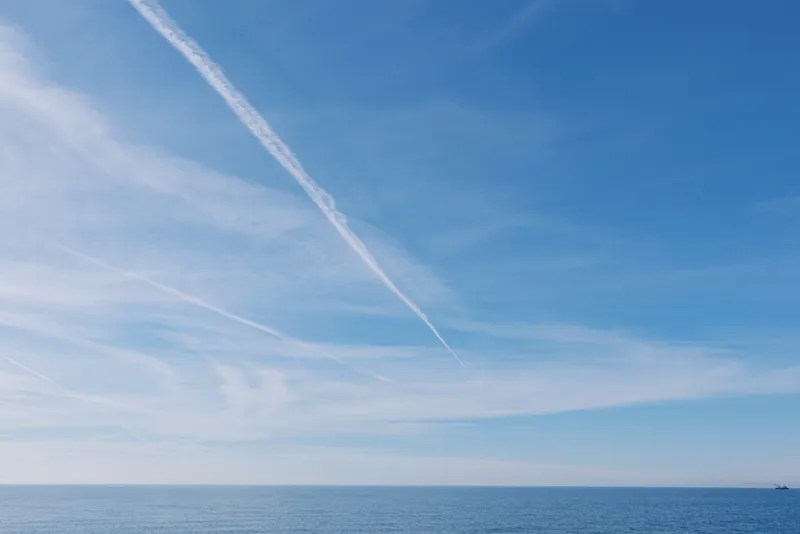 Sky and sea meet at a tranquil horizon with a bright contrail crossing a clear day, capturing blue calm expanse over open waters and vast seascape