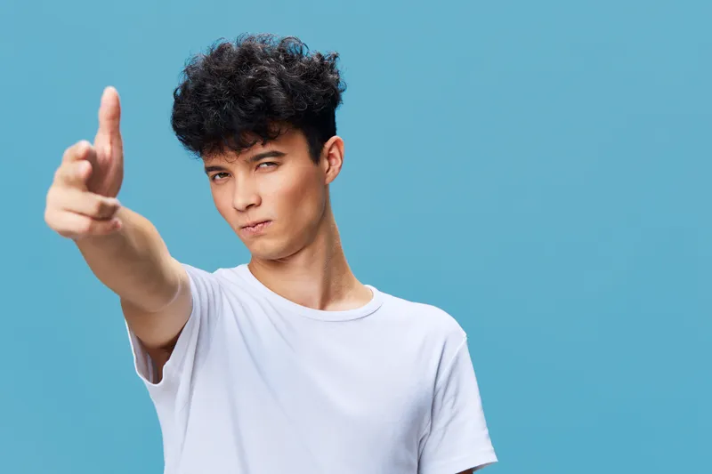 young man pointing finger playfully with serious expression wearing white t-shirt against blue background, isolated portrait with curly hair and casual style