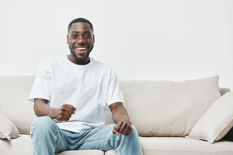 Happy Black man celebrating success while sitting on a sofa, wearing a casual white t shirt, conveying joy and positivity in a minimalistic interior setting
