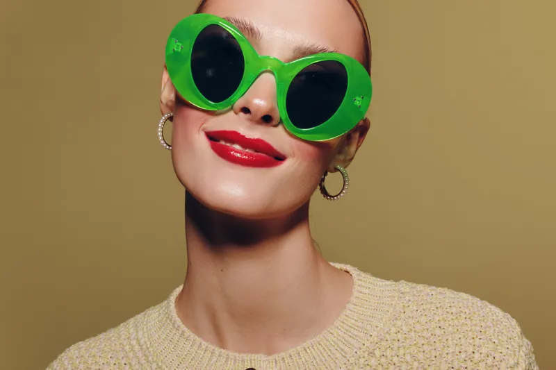 Woman with bright red lipstick wearing large green sunglasses and hoop earrings smiles confidently against beige background. Fashion portrait with bold accessories and stylish makeup concept.