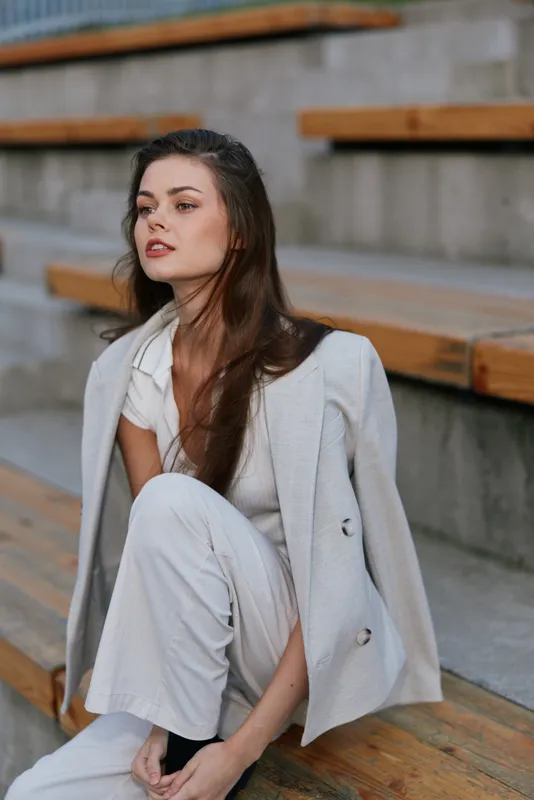Young woman in light suit sitting on wooden steps outdoors with thoughtful expression and natural makeup, casual fashion style in neutral tones