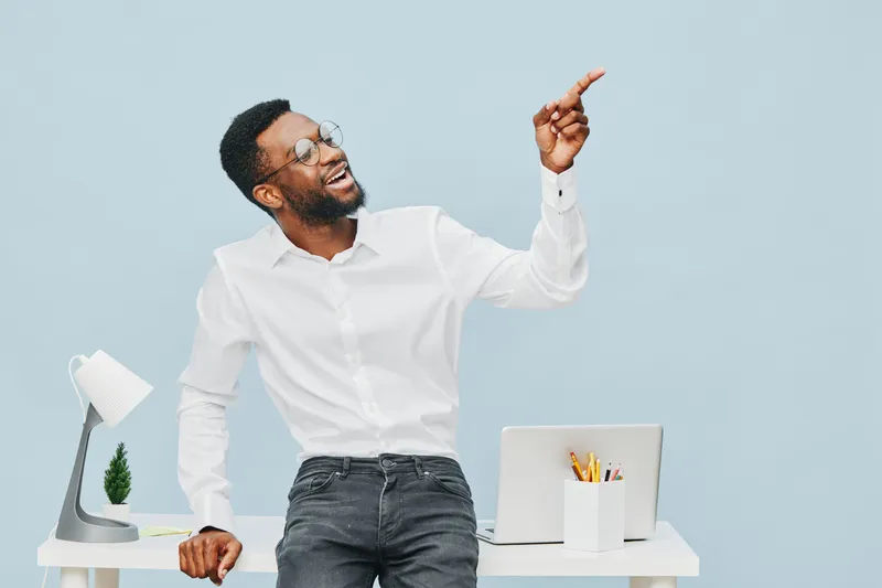 Bright cheerful young African American man with glasses, wearing white shirt and jeans, smiling and pointing upwards while standing at modern workspace with laptop and desk accessories, studio shot. P