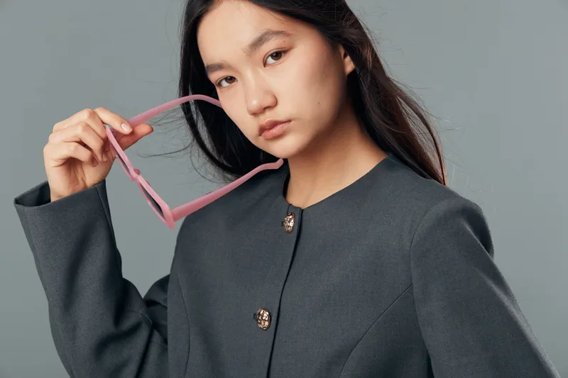 Young woman in dark gray coat holding pink sunglasses with serious expression looking at camera on gray background for fashion and lifestyle portrait studio.