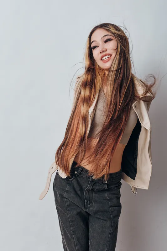Young woman with long brown hair posing against white background wearing casual jacket and black jeans, smiling and expressing carefree mood in studio portrait.