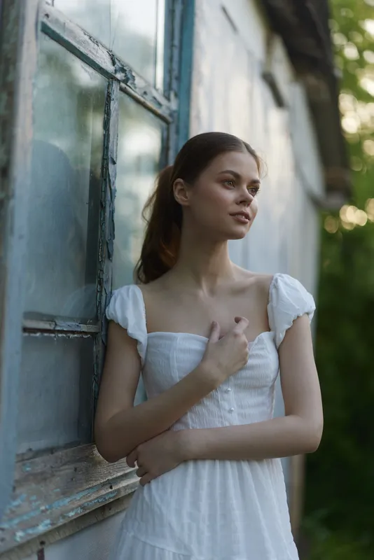 Elegant young woman in a white dress standing by a rustic window, exuding a serene and thoughtful emotion in a lush green outdoor setting