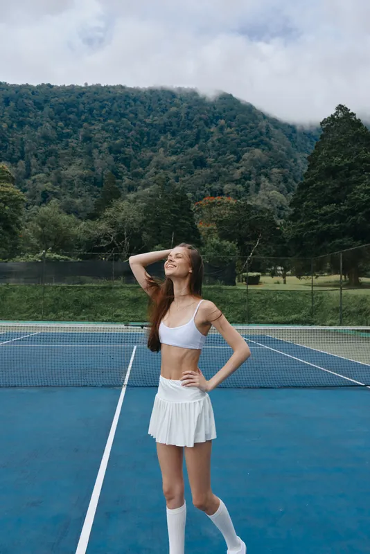 Young athletic woman in a white tennis outfit, smiling joyfully on a tennis court surrounded by lush greenery, portraying an energetic and healthy lifestyle