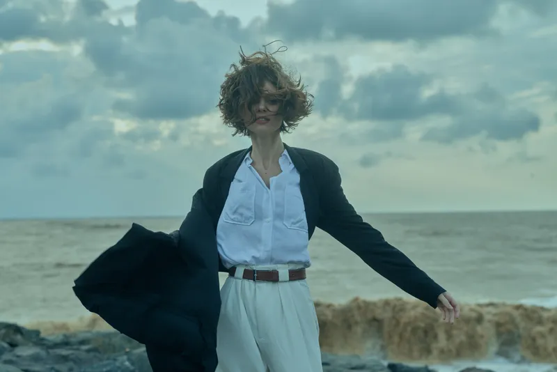 Female professional in a blazer and white shirt on a windy beach, holding her coat open as waves crash, capturing bold fashion and coastal movement against a vast ocean backdrop