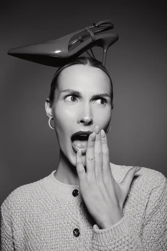 Surprised Caucasian woman with light skin wearing textured sweater and high heel shoe on head expresses shock with hand near mouth in monochrome studio.