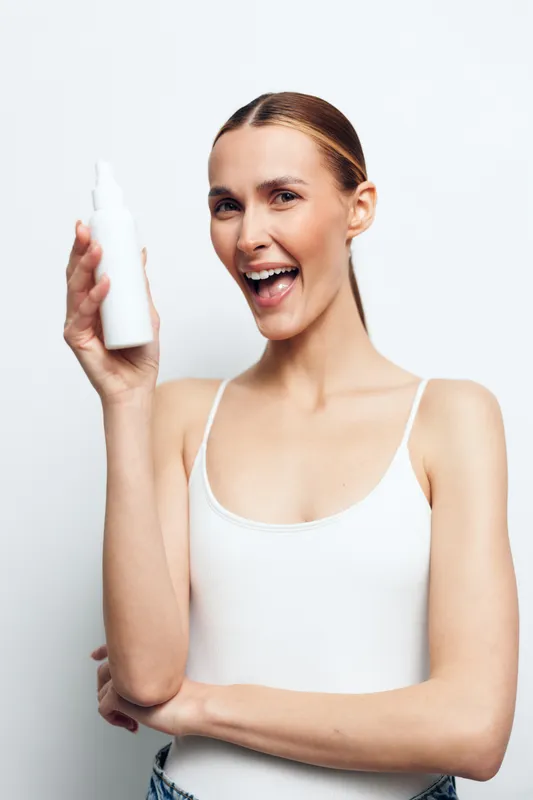 Cheerful woman holding a bottle, showcasing skincare product, bright studio lighting, clean white background, casual outfit, confident mood, natural tones, product focus