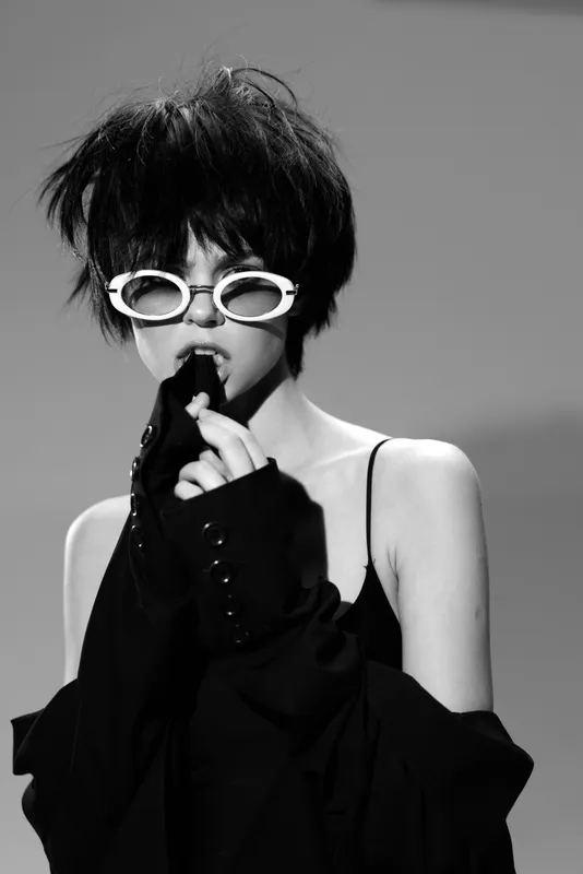 Black and white portrait of a stylish woman wearing white sunglasses and dark clothing with a moody expression and edgy hairstyle.