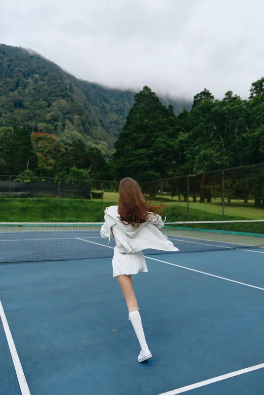 Young woman celebrating on a tennis court in a white outfit, surrounded by lush green mountains and a cloudy sky, showcasing joy and freedom while enjoying sports
