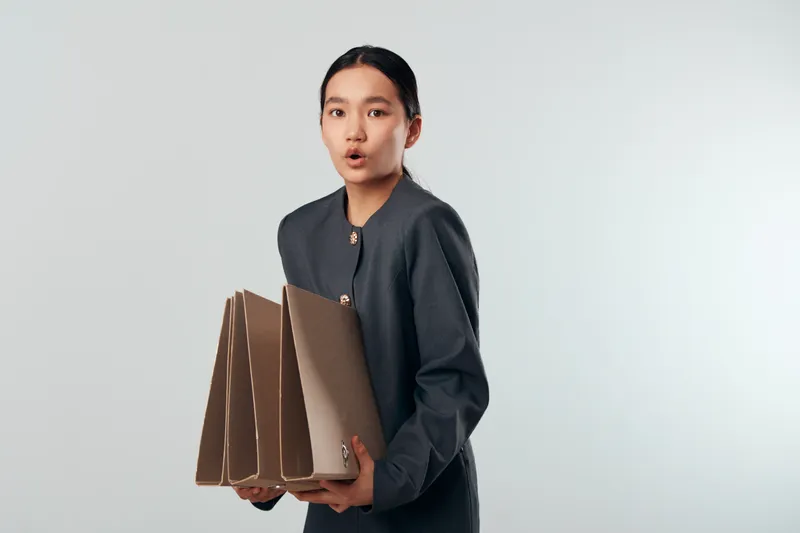 Young woman holding multiple file folders with surprised expression standing against plain background for office work, document organization and business tasks.