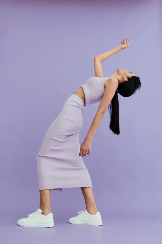 woman posing in lilac outfit stretches backward gracefully against soft purple studio background, showcasing elegance and modern fashion style with white sneakers