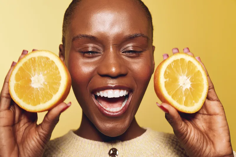 Happy African woman with dark skin holds two orange halves near her face, smiling with eyes closed against a yellow background. Concept of healthy lifestyle and fresh fruit.