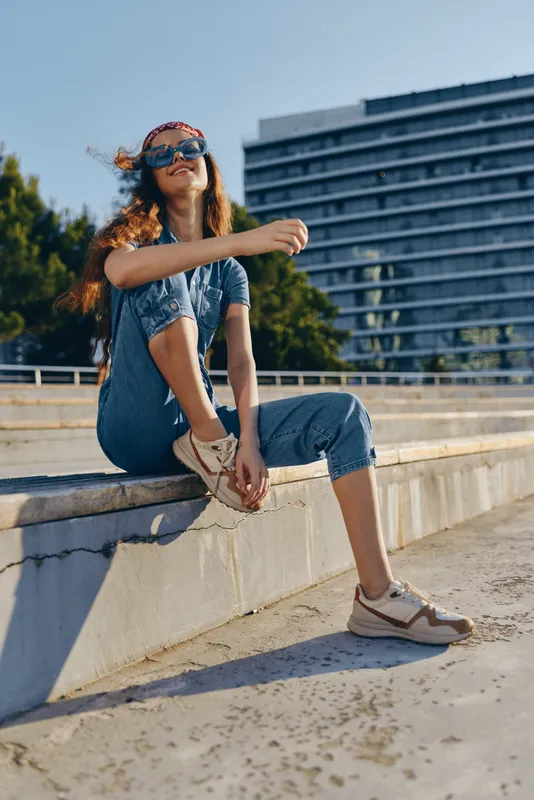 smiling woman sitting outdoors in casual denim jumpsuit and sneakers with sunglasses and headband on a sunny day near modern building