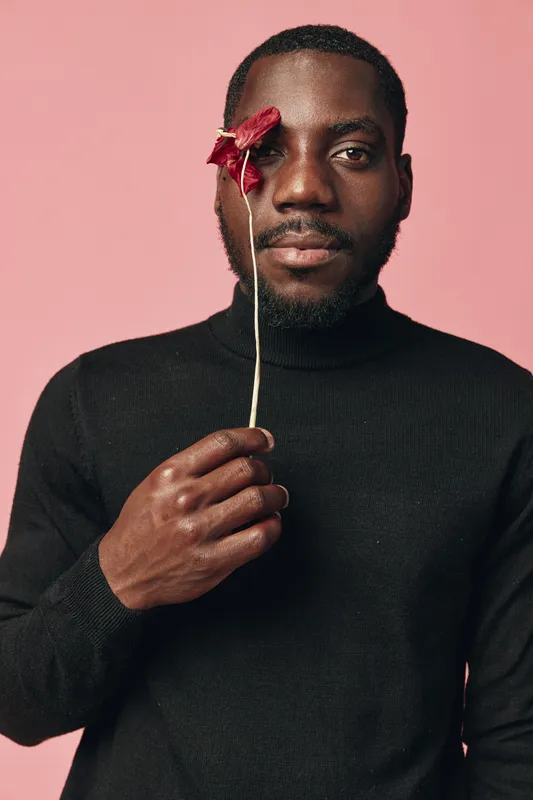 African man with dark skin holding red flower near eye in creative conceptual visual style. Portrait with pink background and black sweater for artistic expression and emotion.