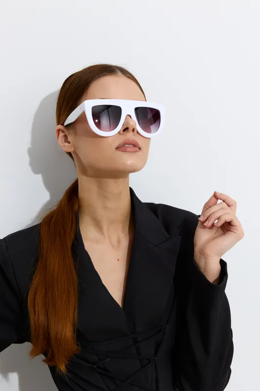 Fashion woman with long red hair wearing oversized white sunglasses and a black blazer posing against a white background in a modern stylish look.