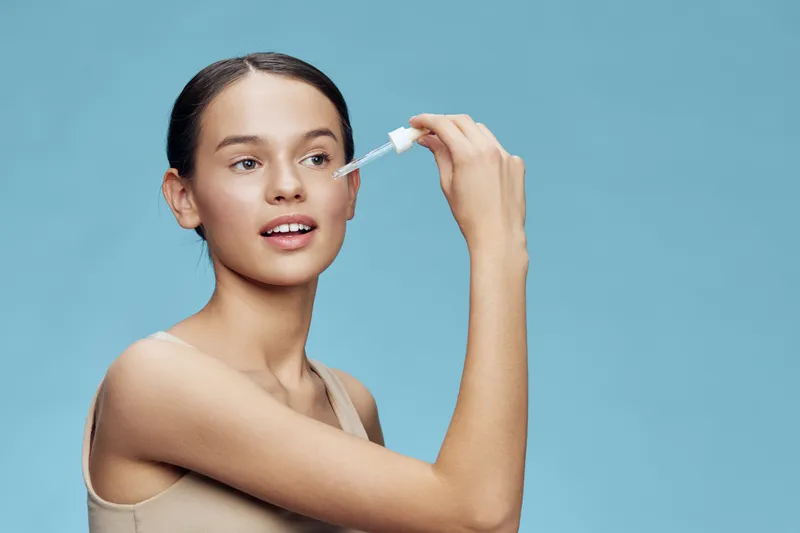 Young woman applying facial serum with dropper against a light blue background, showcasing healthy skincare routine and glowing skin