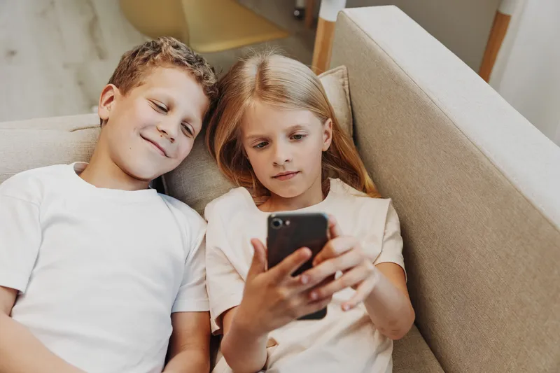 Two children lying on a couch looking at a smartphone together, sharing a moment of curiosity and connection in a cozy home environment.